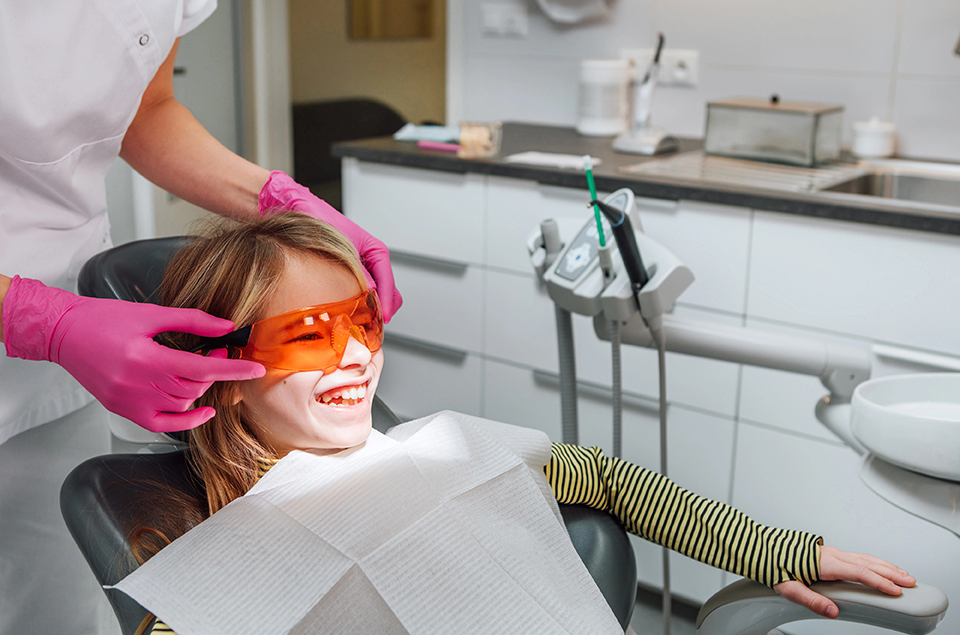 Dentist doctor wearing protective eyeglasses to little girl during teeth restoration, dental fillings procedure with curing UV light. Health care, teeth beauty and medicare industry concept image,