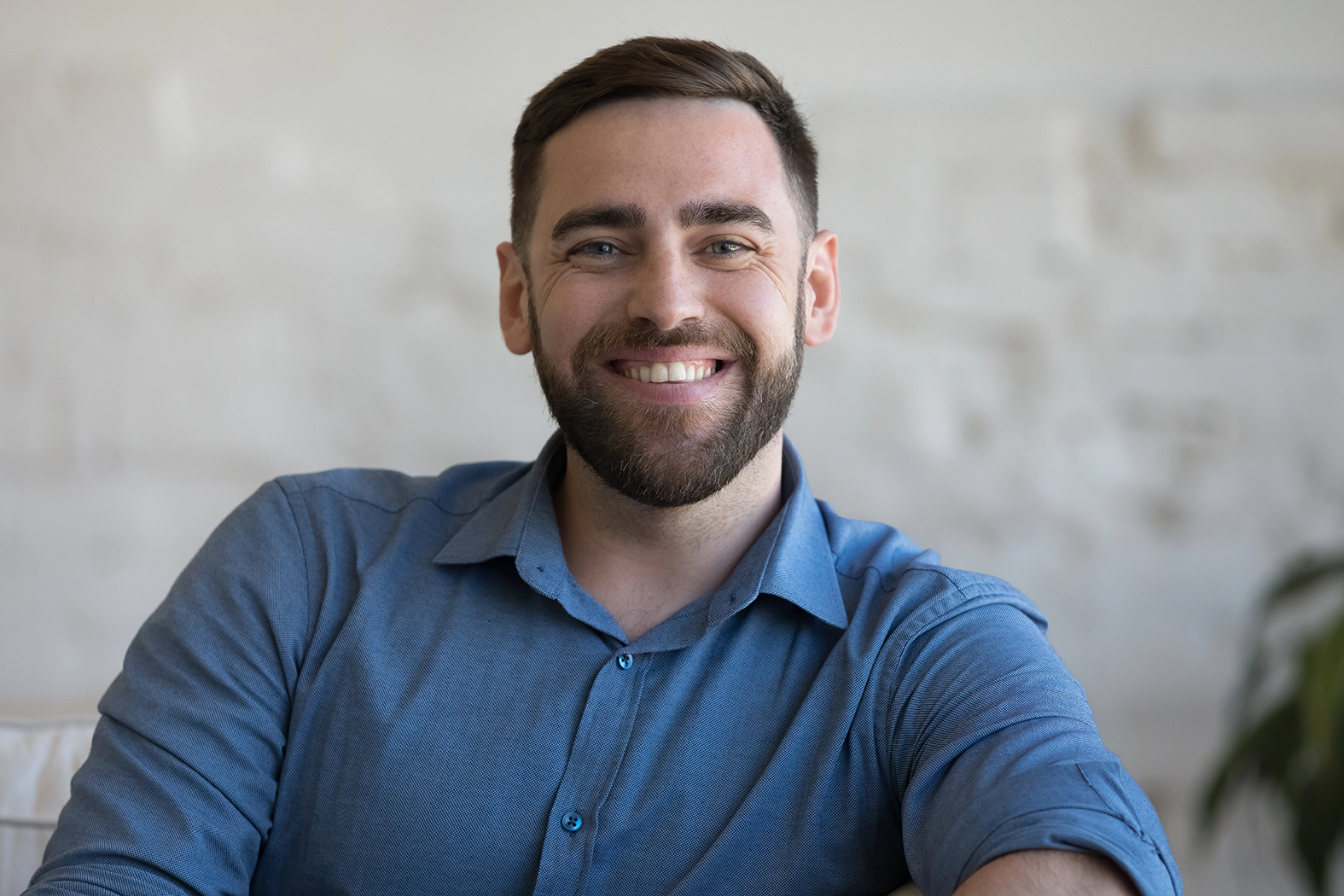 Cheerful confident handsome adult man sitting on couch, armchair at home against white wall background, looking at camera, smiling. Happy homeowner, blogger, freelancer head shot portrait