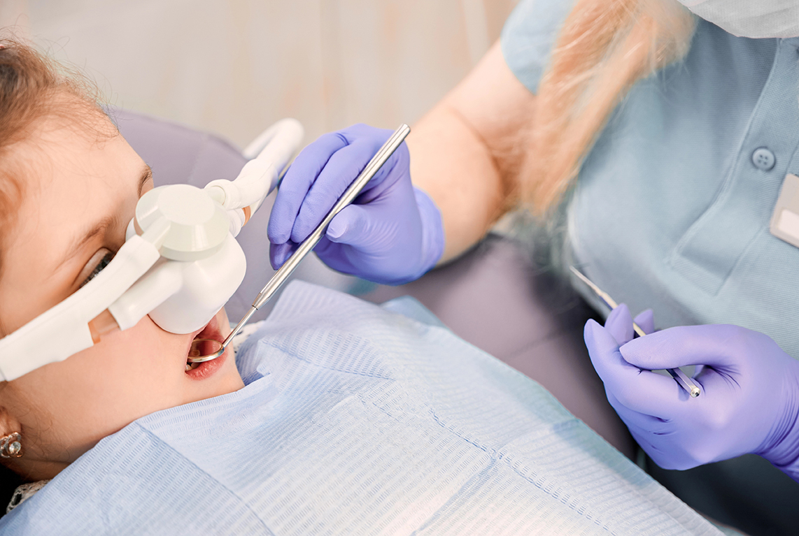 Female dentist checking child teeth with dental explorer and mirror while girl lying in dental chair with inhalation sedation. Concept of pediatric, sedation dentistry and dental care.